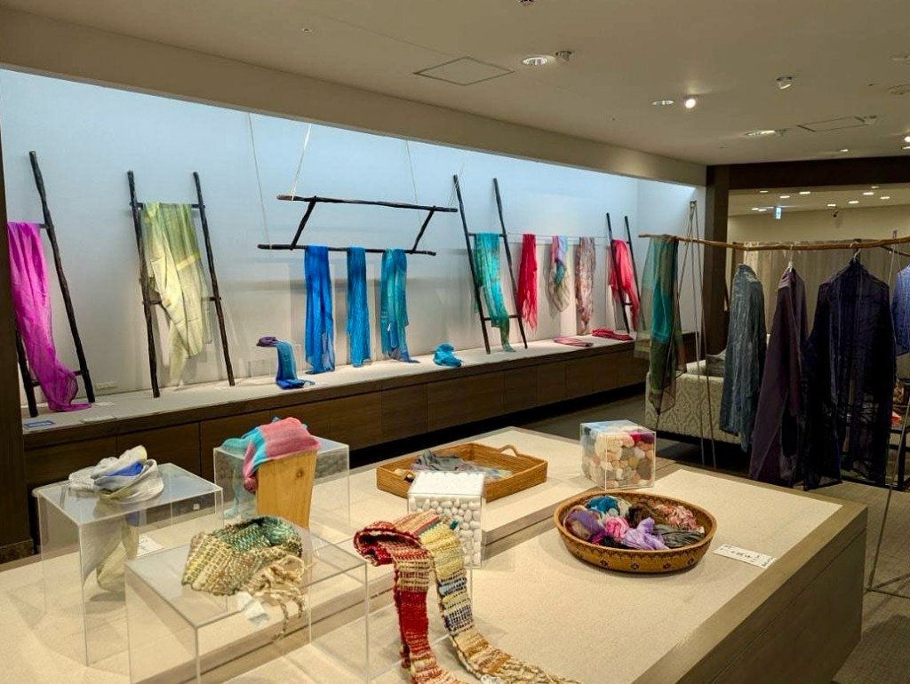色とりどりの布が展示された広島三越のアートギャラリーの内部。手前のテーブルには糸や布のサンプルが並べられ、背後には吊るされた染められたストールや布が見える。