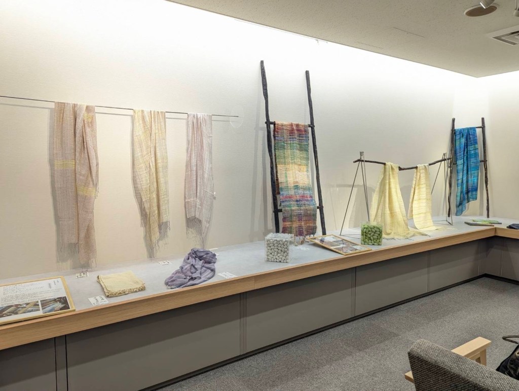 A display of various textile pieces and scarves showcased on wooden frames at the Fukuyama Tenmaya Art Gallery, highlighting colors and textures.