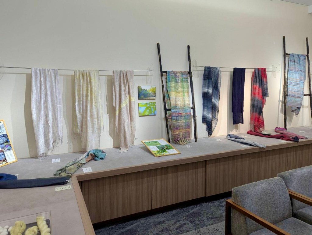 展示されたさまざまな色とテクスチャの布が壁に掛けられ、手前にはテーブルに置かれた布が展示されているアートギャラリーの風景。