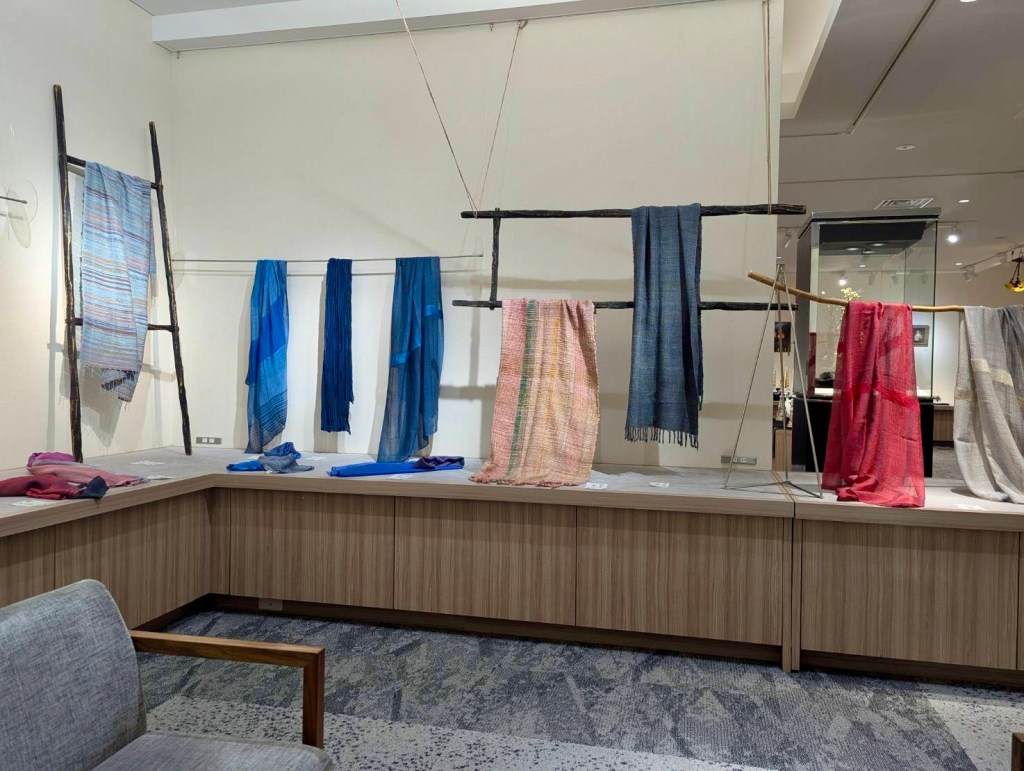 An art gallery display featuring a variety of colorful handwoven textiles hanging on racks, with a wooden table underneath displaying more fabrics.